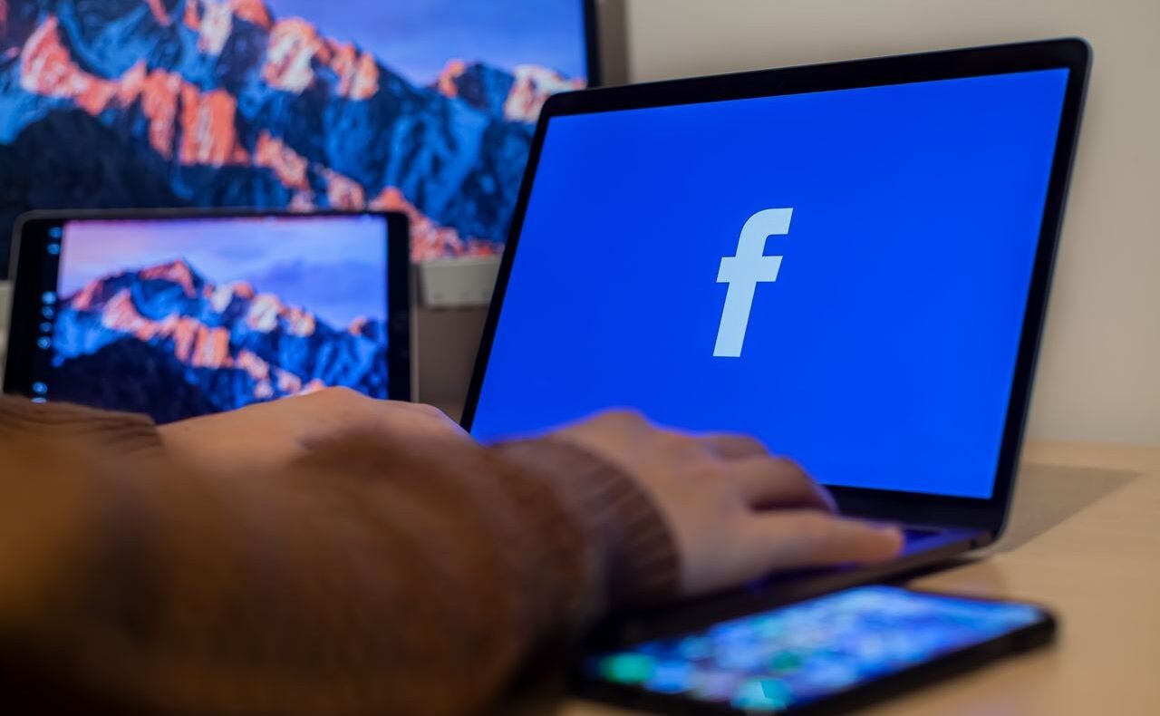 Laptop displaying Facebook at workspace with multiple devices, highlighting modern technology.